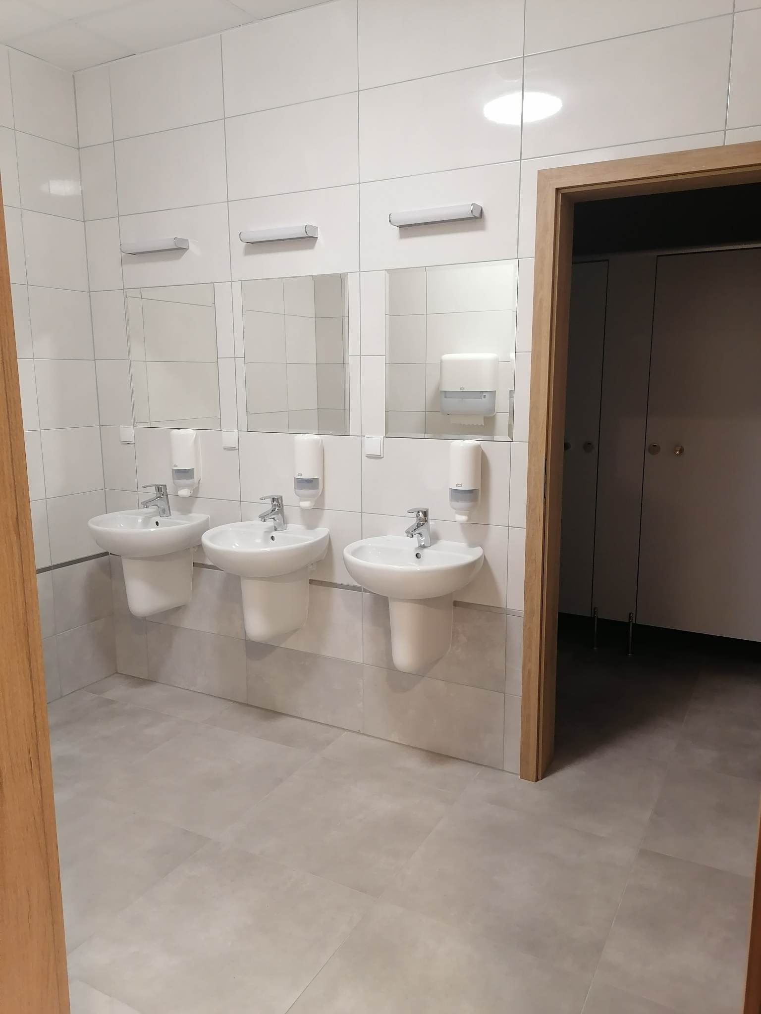 Publiczna toaleta z trzema umywalkami, białymi płytkami na ścianach, lustrami i dozownikami mydła, widok z perspektywy drzwi.