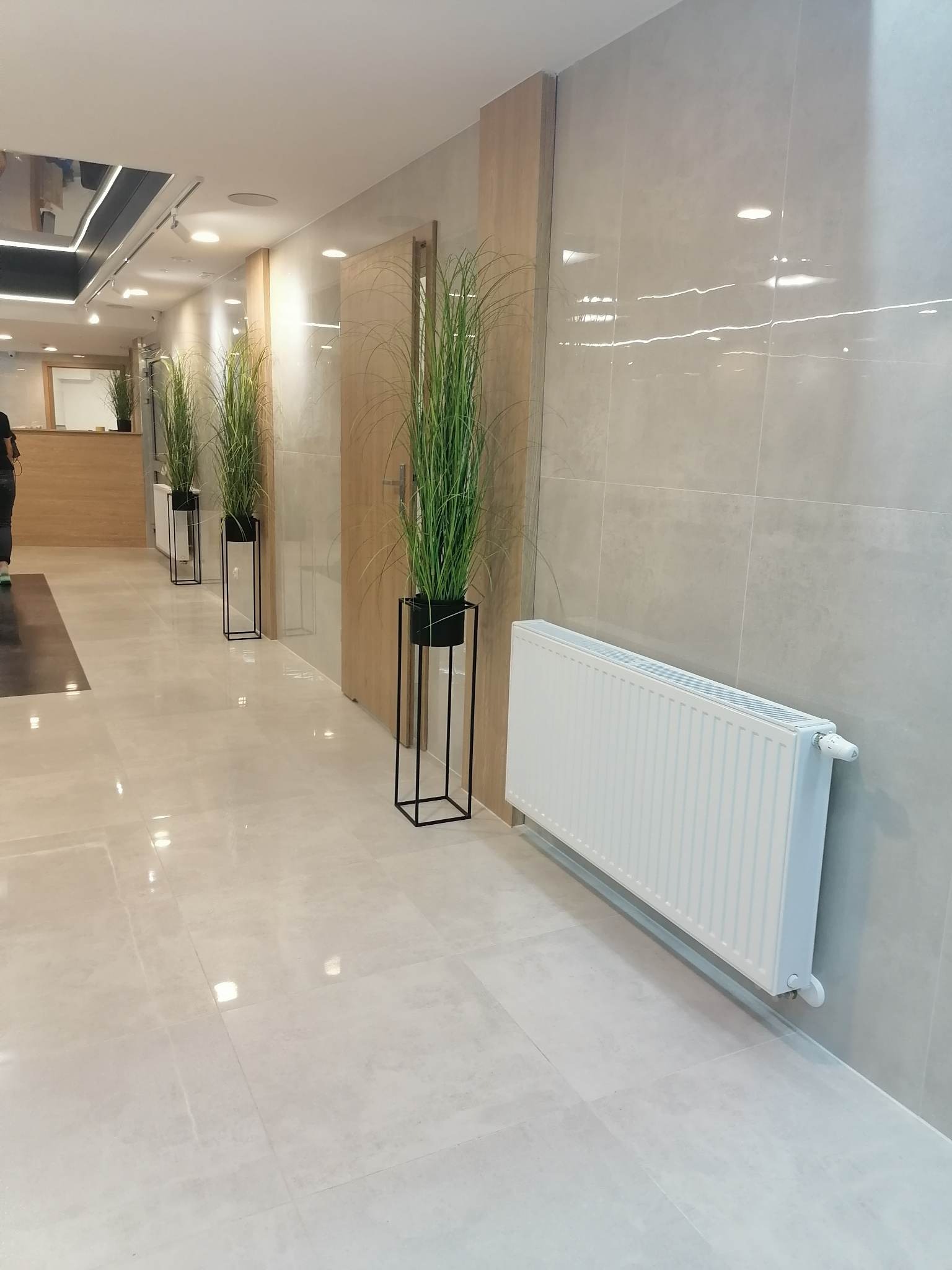 Jasne, błyszczące płytki podłogowe i ścienne w korytarzu, z wysokimi roślinami w minimalistycznych donicach i białym grzejnikiem panelowym.