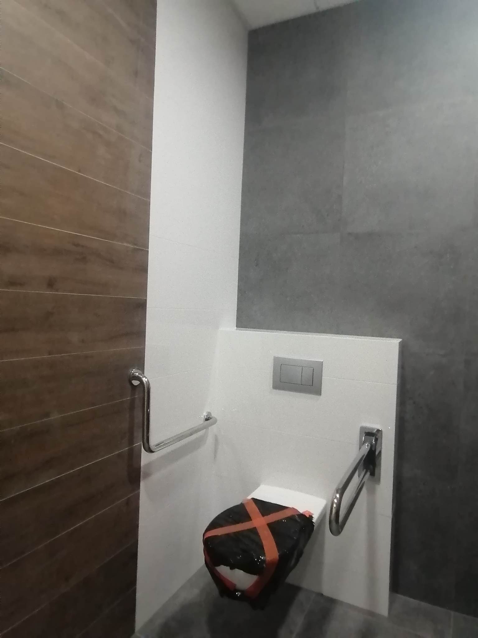 Nowoczesna łazienka w trakcie remontu: brązowe płytki imitujące drewno, szare płytki na ścianie, biała zabudowa toalety z metalowymi uchwytami dla niepełnosprawnych, sedes zabezpieczony czarną...