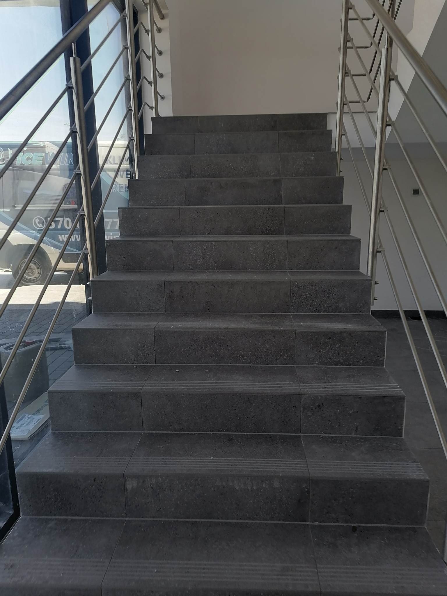 Schody wyłożone ciemnoszarą, matową płytką z metalową balustradą, widok z dołu na górę, z widocznym fragmentem okna i samochodu na zewnątrz.