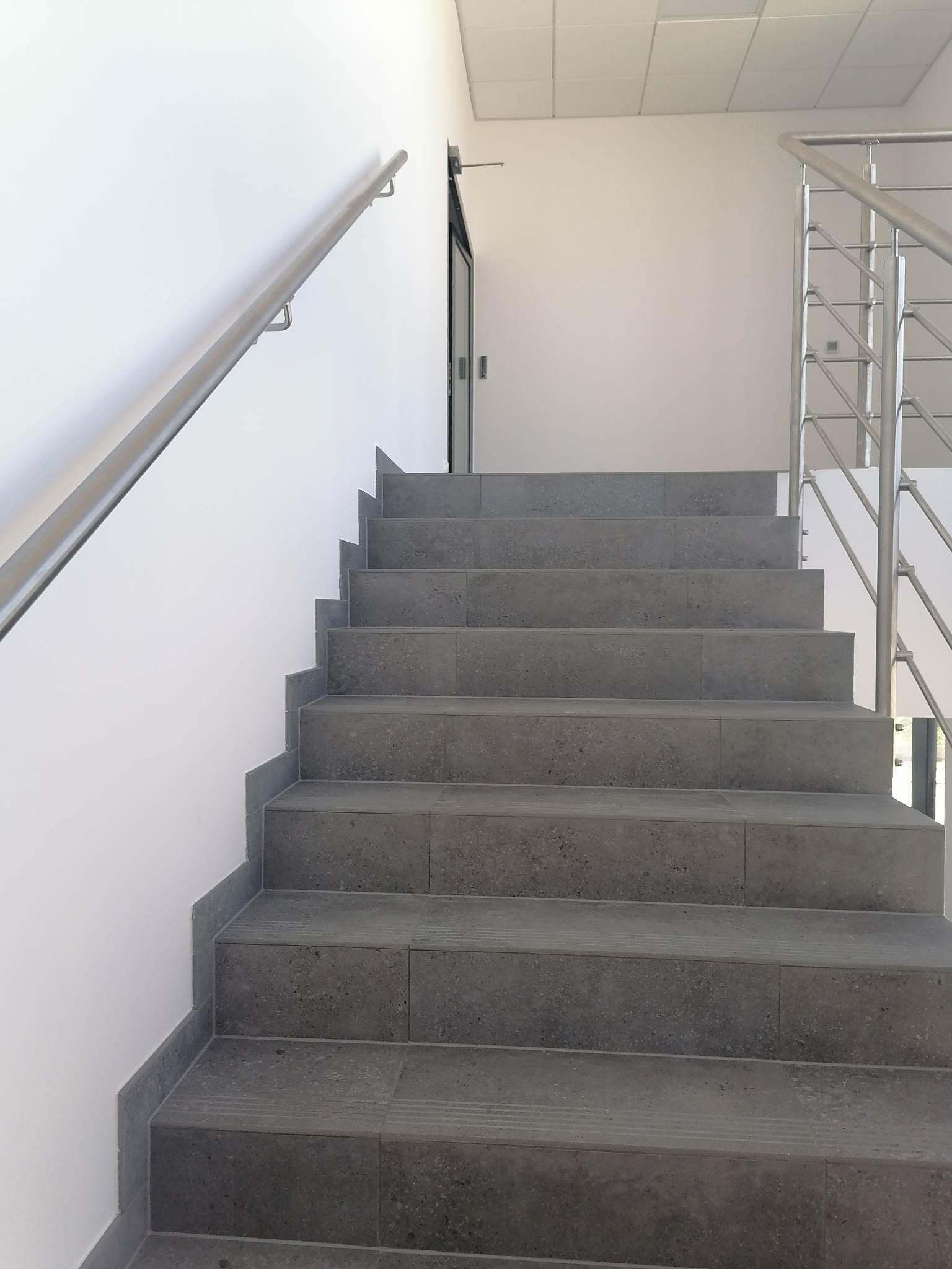 Schody wyłożone szarą, kamienną płytką z metalową balustradą i poręczą, prowadzące do góry w nowoczesnym wnętrzu.