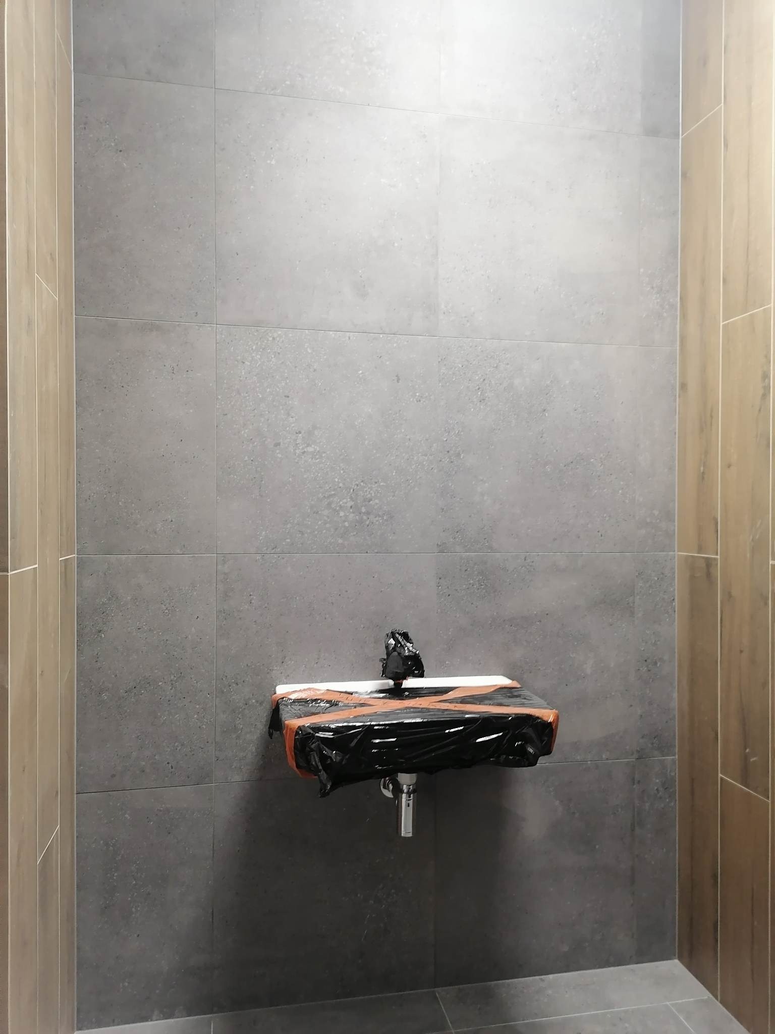 Ściana łazienki wyłożona szarymi, matowymi płytkami wielkoformatowymi, umywalka zabezpieczona folią ochronną w trakcie prac remontowych, widoczne pionowe wykończenia ściany imitujące drewno.