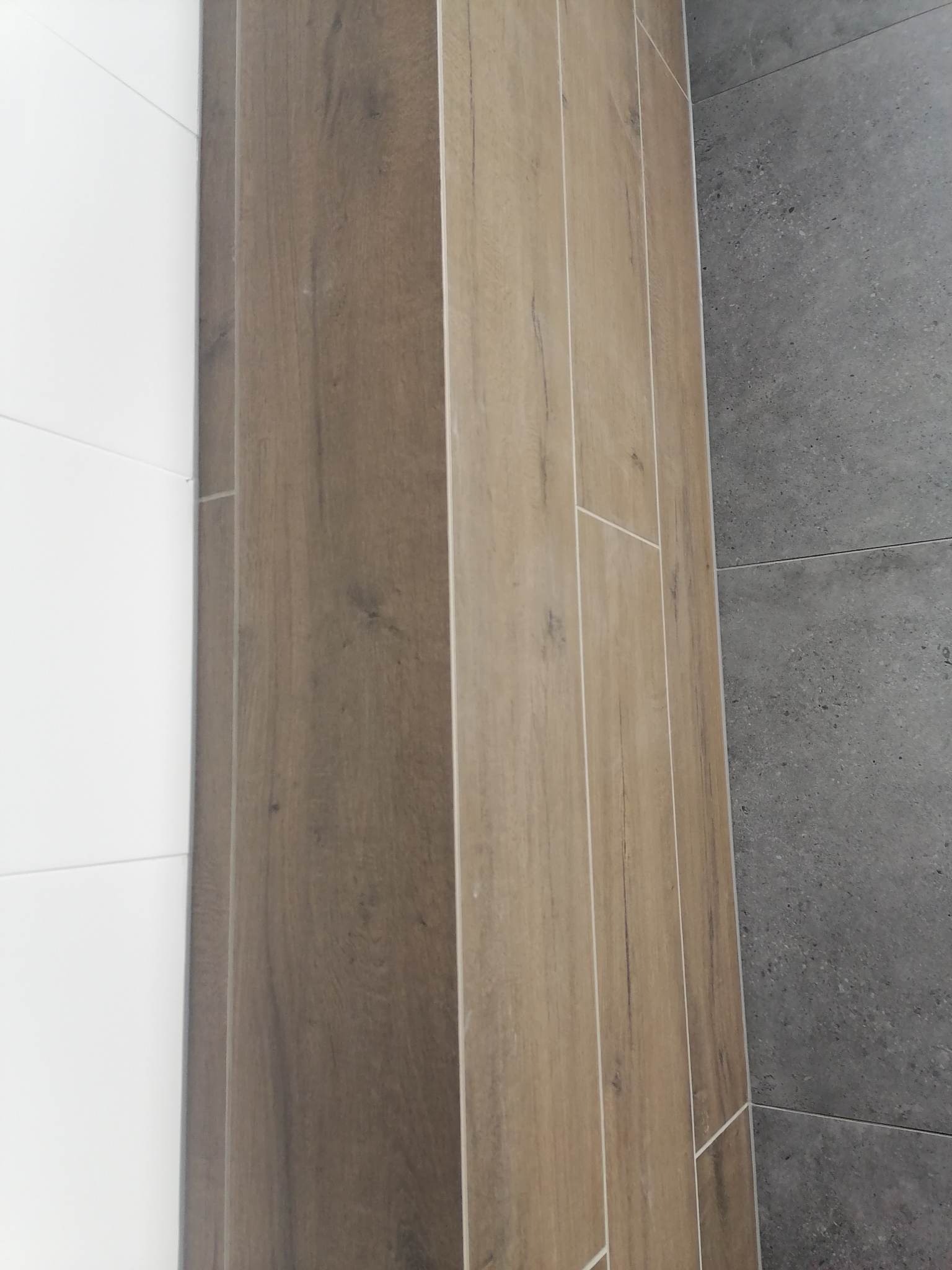 Ułożone pionowo płytki imitujące drewno w odcieniach brązu sąsiadują z białymi płytkami po lewej i szarymi płytkami o teksturze betonu po prawej stronie.