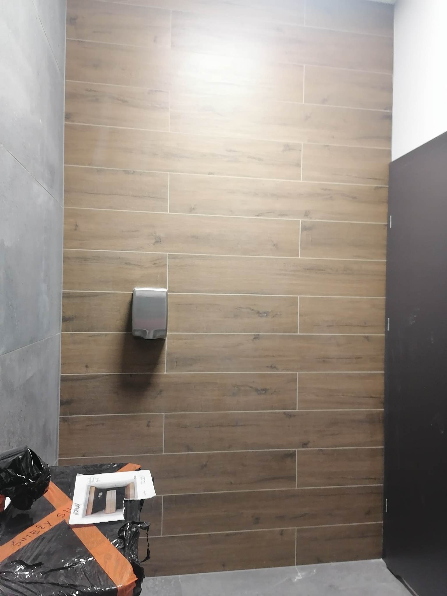 Ukończona łazienka z płytkami imitującymi drewno na ścianie, szara ściana po lewej, czarne drzwi po prawej, dozownik do mydła na ścianie, na podłodze zabezpieczone folią śmieci.