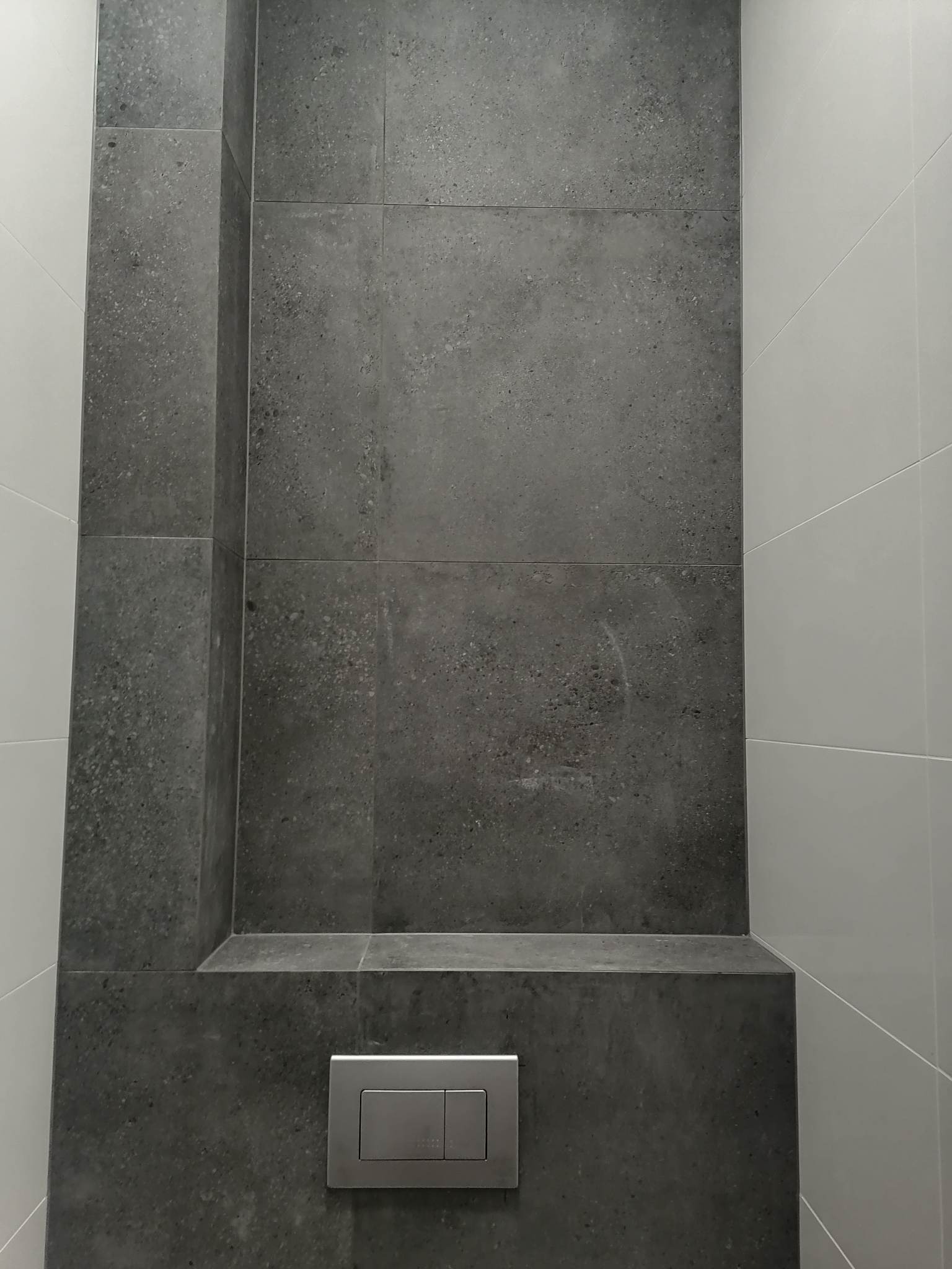 Szare, matowe płytki imitujące kamień, ułożone w zabudowie geberit w łazience w Toruniu, kontrastują z białymi, błyszczącymi płytkami na sąsiedniej ścianie. Widoczny metalowy panel spłuczki.