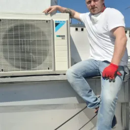 Mężczyzna w czerwonych rękawicach opiera się o zewnętrzną jednostkę klimatyzacji Daikin na dachu, z maską na szyi, w tle błękitne niebo.