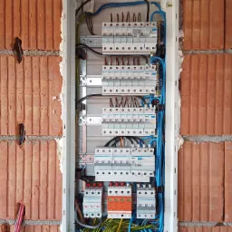 Rozdzielnica elektryczna Hager zamontowana w surowej ścianie z czerwonej cegły, widoczne kable w kolorach niebieskim, czarnym, brązowym, żółto-zielonym oraz zabezpieczenia nadprądowe...