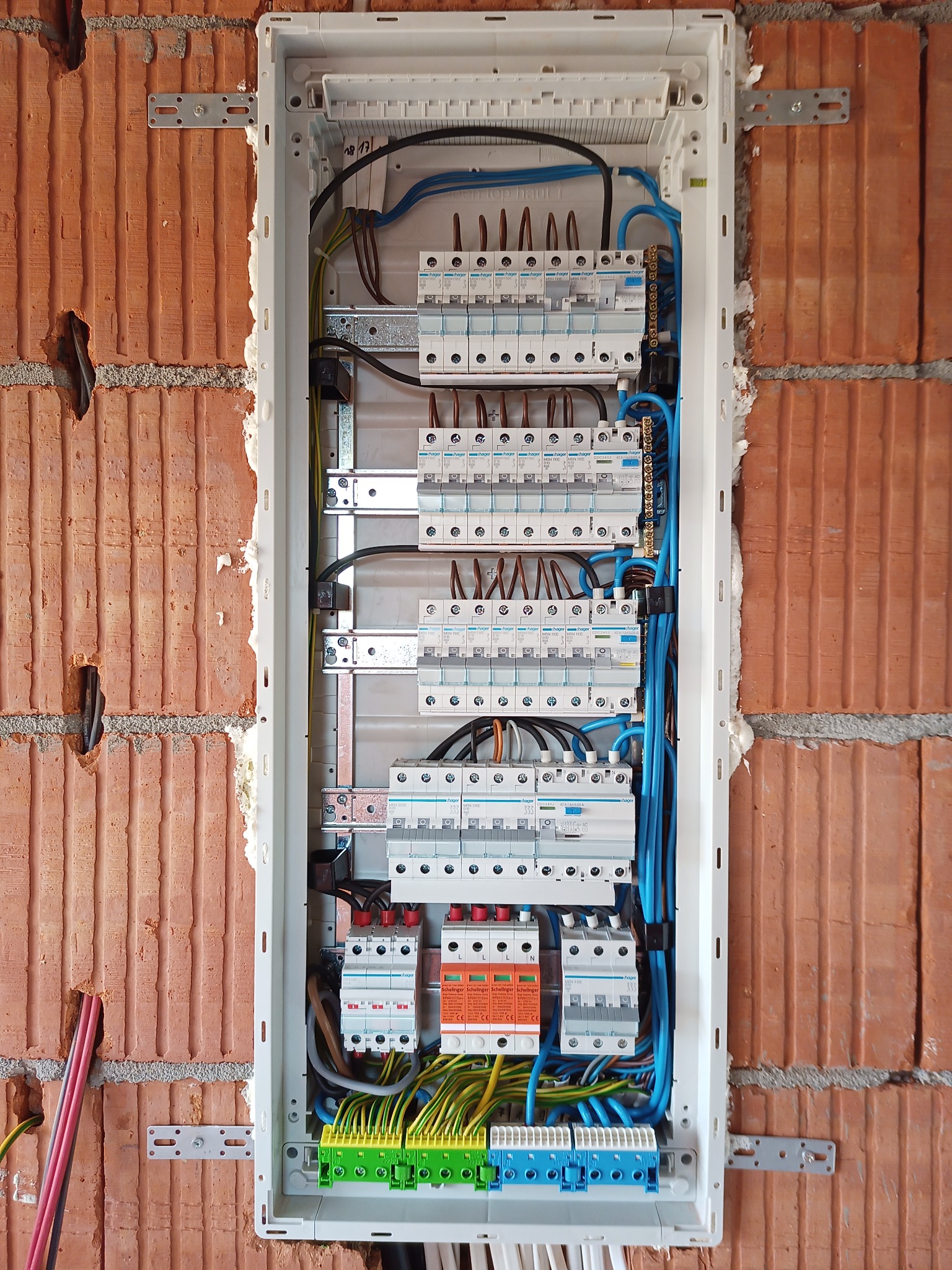 Rozdzielnica elektryczna Hager zamontowana w surowej ścianie z czerwonej cegły, widoczne kable w kolorach niebieskim, czarnym, brązowym, żółto-zielonym oraz zabezpieczenia nadprądowe...