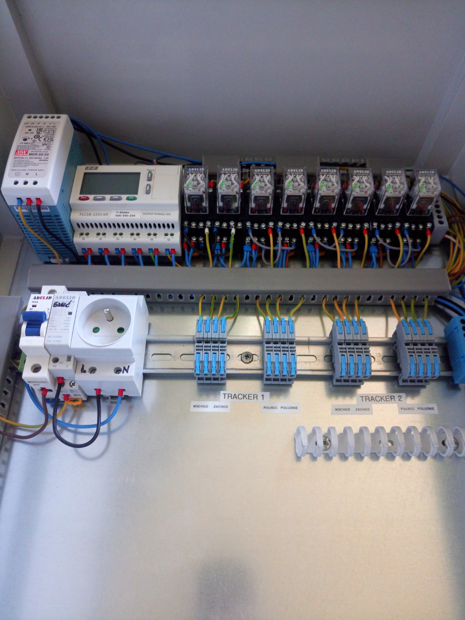 Wnętrze metalowej szafy sterowniczej z podzespołami elektrycznymi: zasilacz, sterownik PLC, przekaźniki, gniazdo z bezpiecznikiem, listwy zaciskowe z opisami Tracker 1 i Tracker 2.