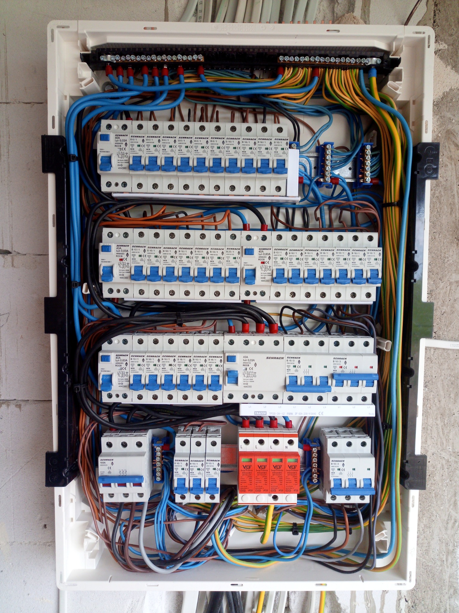 Otwarta skrzynka rozdzielcza z licznymi bezpiecznikami Schrack, przewodami w różnych kolorach i modułami przeciwprzepięciowymi Vcx, zamontowana na surowej ścianie z betonu komórkowego.
