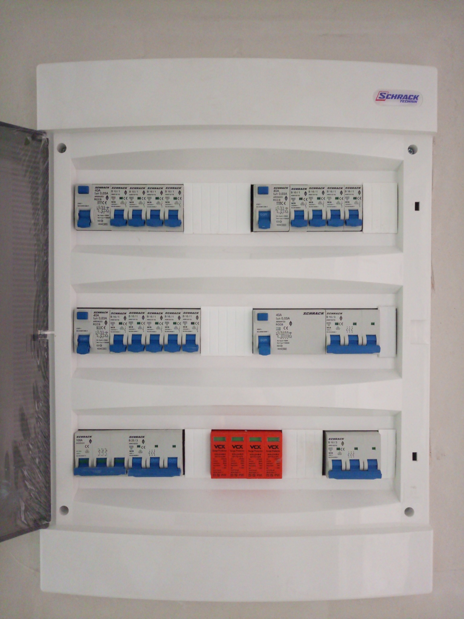 Otwarta skrzynka elektryczna Schrack z zainstalowanymi bezpiecznikami i wyłącznikami różnicowoprądowymi, widoczne zabezpieczenia przeciwprzepięciowe VCX, zamontowana na ścianie.