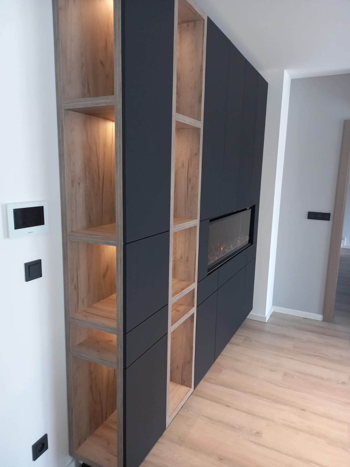 Ciemnoszara zabudowa meblowa z drewnianymi półkami i elektrycznym kominkiem, oświetlenie LED w półkach, widok z boku, podłoga z paneli.