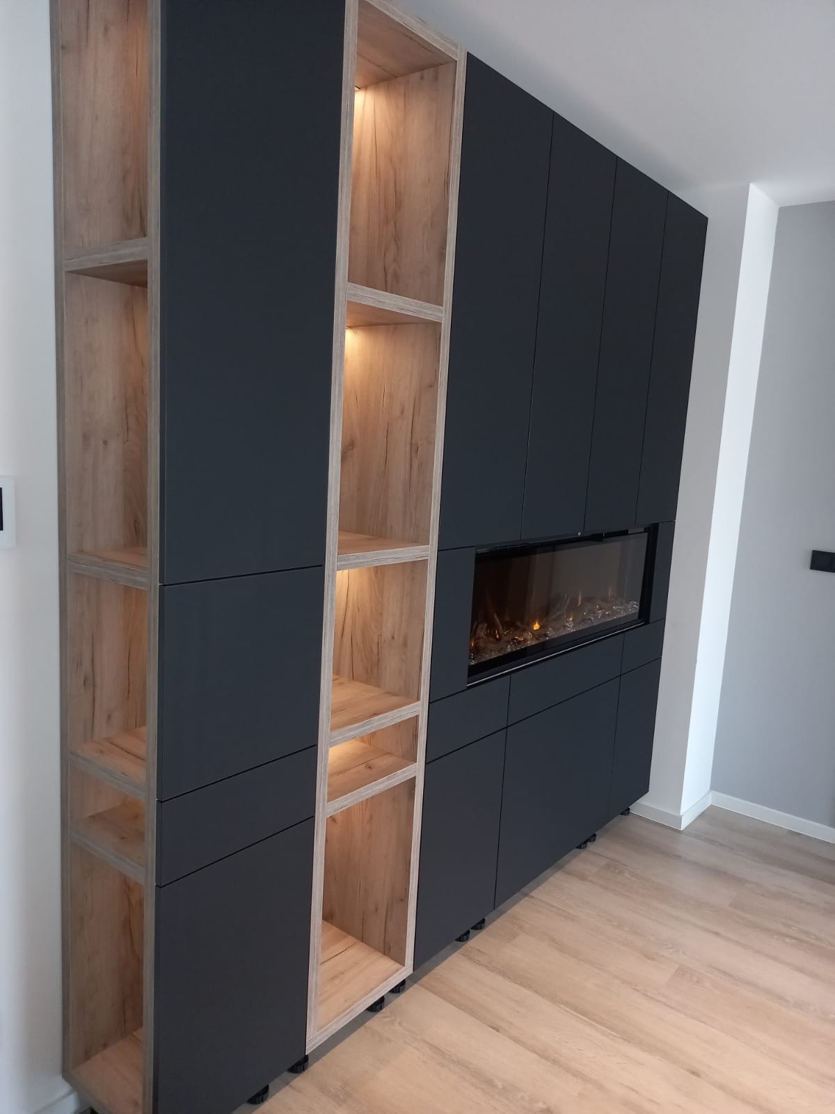 Ciemnografitowa zabudowa meblowa z drewnianymi wnękami, podświetlonymi od wewnątrz, z wkomponowanym elektrycznym kominkiem.