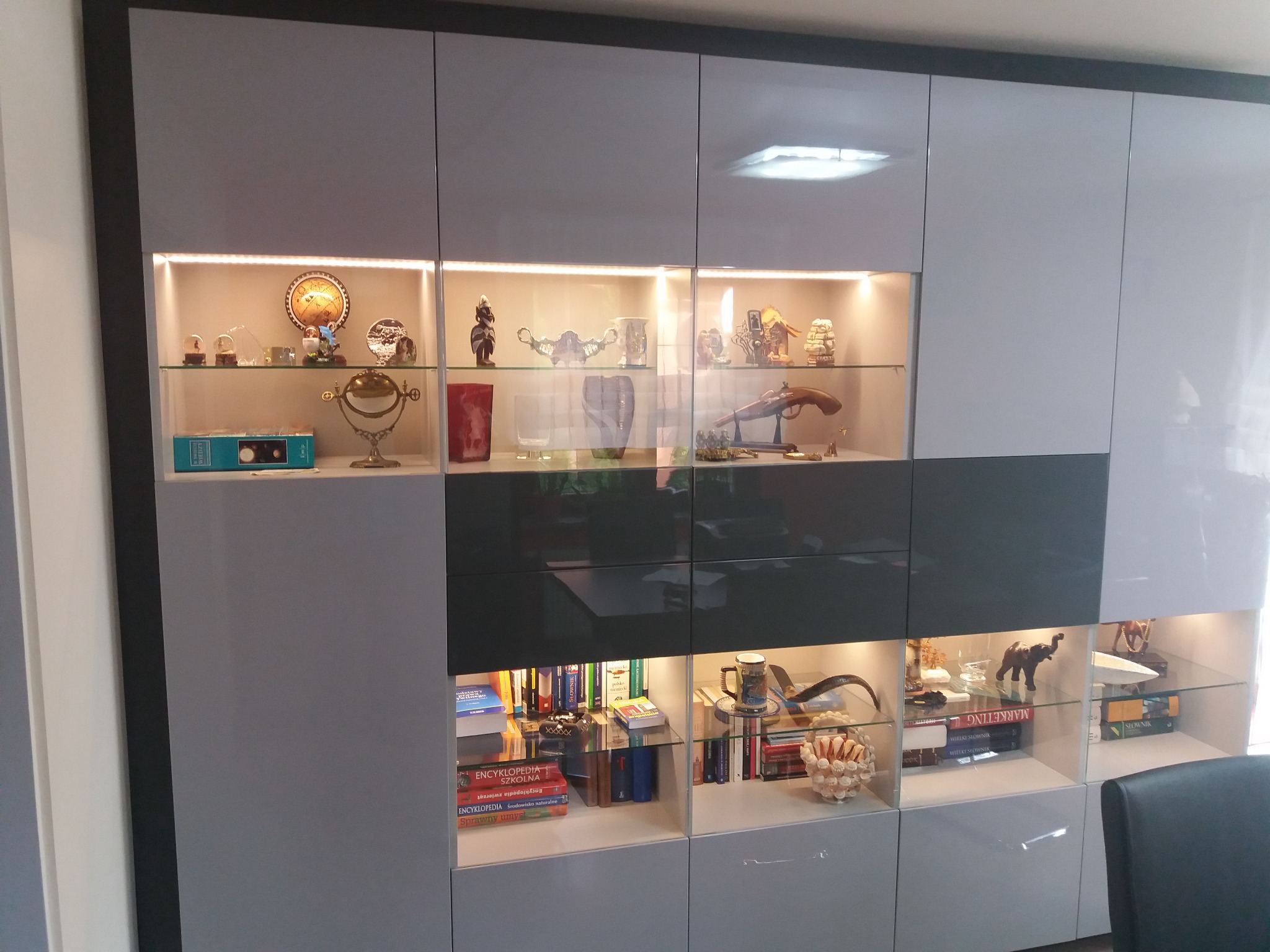 Duża, nowoczesna szafa z szarymi i białymi frontami, z oświetlonymi półkami szklanymi, na których znajdują się książki, ozdoby i figurki dekoracyjne.