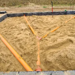 Fundament otoczony piaskiem z widocznym systemem rur drenarskich w kolorze pomarańczowym, czarne obrzeże fundamentu, w tle ziemia.