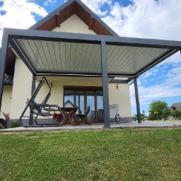 Nowoczesna, szara pergola z jasnym zadaszeniem lamelowym, przylegająca do jasnego domu z brązowymi oknami, na pierwszym planie trawnik.