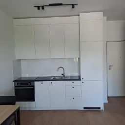 Minimalistyczna biała zabudowa kuchenna z czarnym blatem i płytą grzewczą, widok na szafki g&oacute;rne i dolne, oraz fragment stołu z krzesłem.