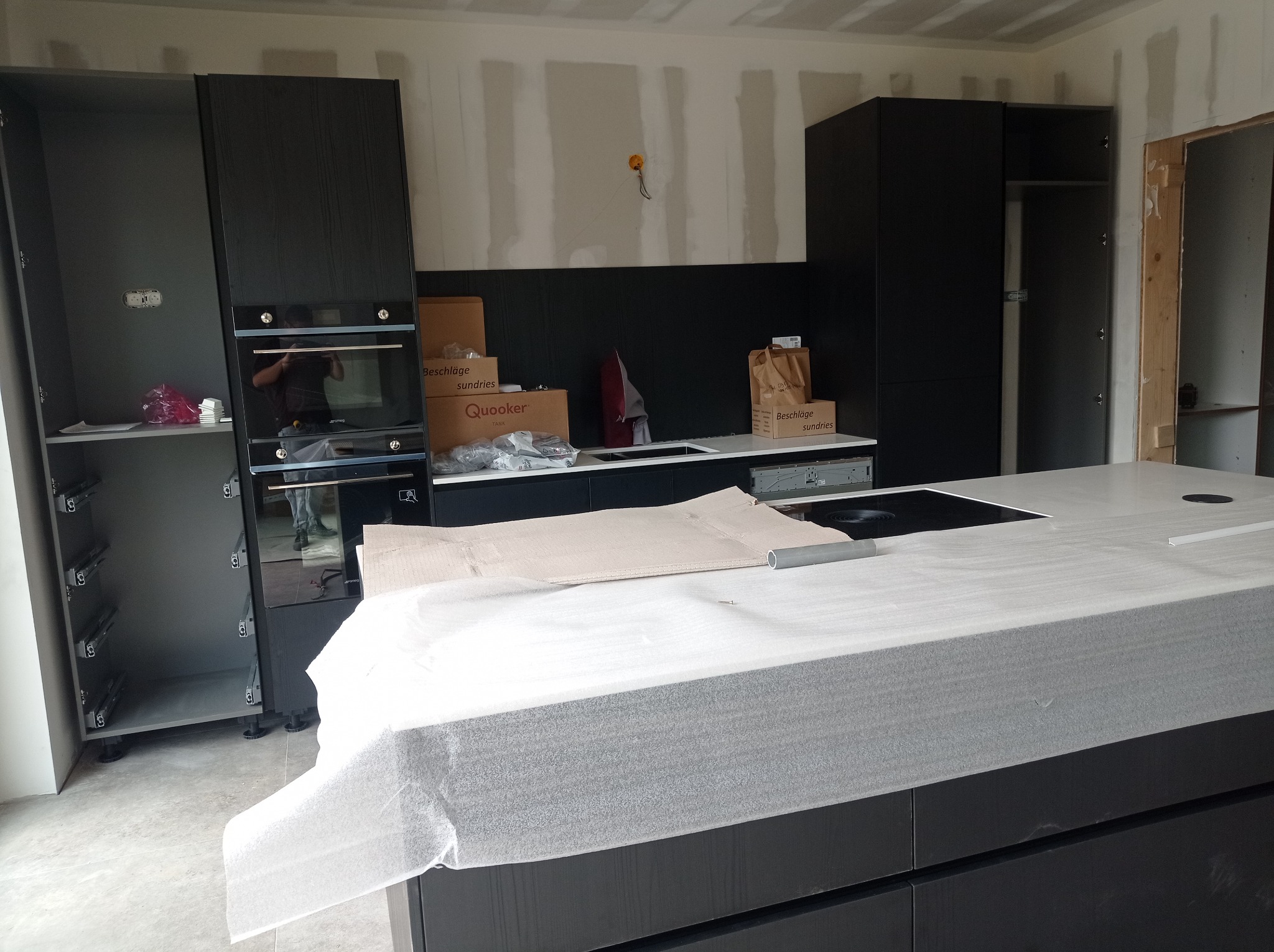 Montaż czarnej kuchni: wyspa z blatem zabezpieczonym folią, zabudowa z piekarnikiem Smeg i otwartą szafką, kartony z akcesoriami w tle.