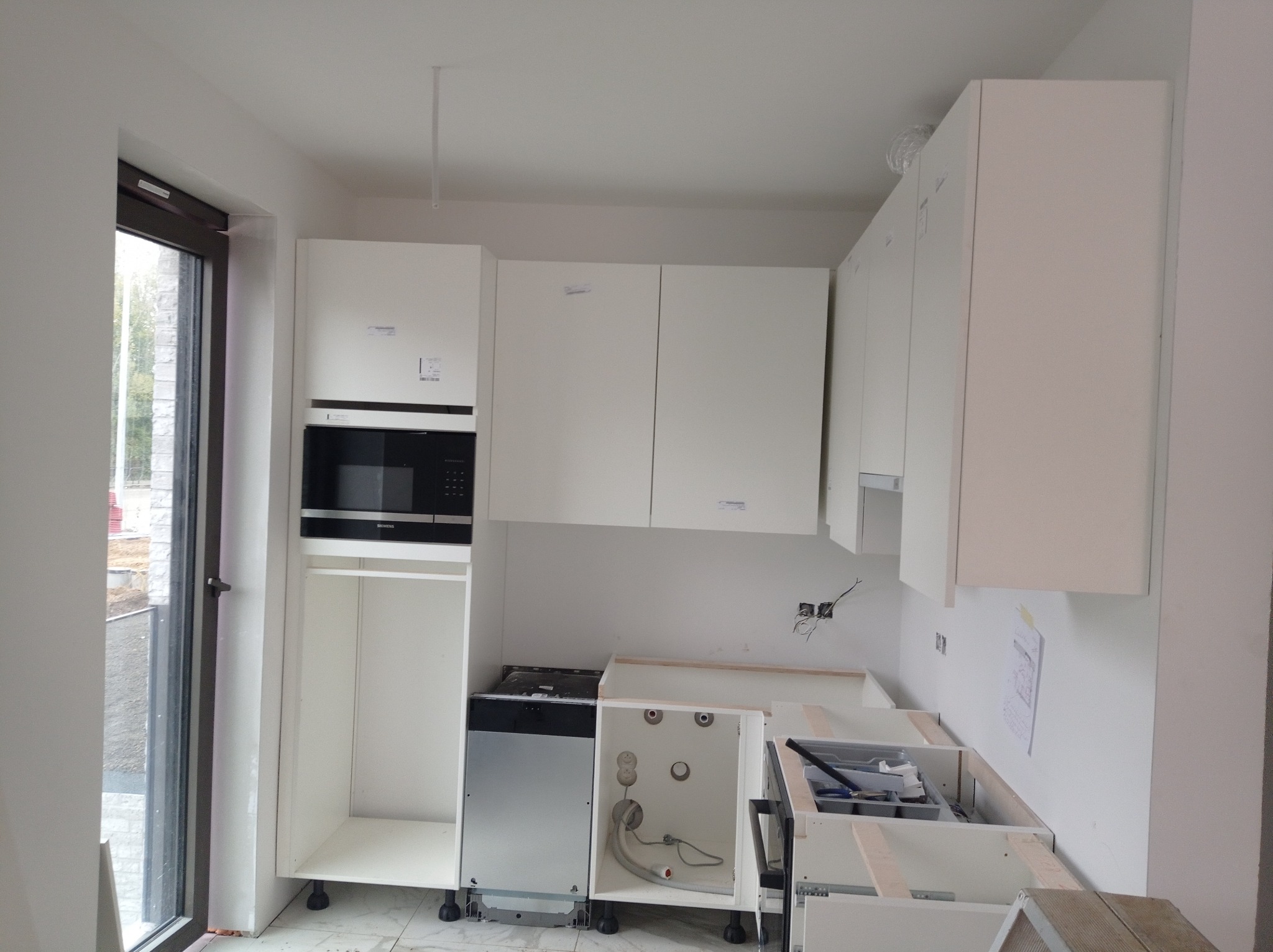 Montaż białej zabudowy kuchennej z wbudowanym piekarnikiem i zmywarką, widoczne niedokończone szafki dolne z instalacją hydrauliczną, okno z widokiem na plac budowy.