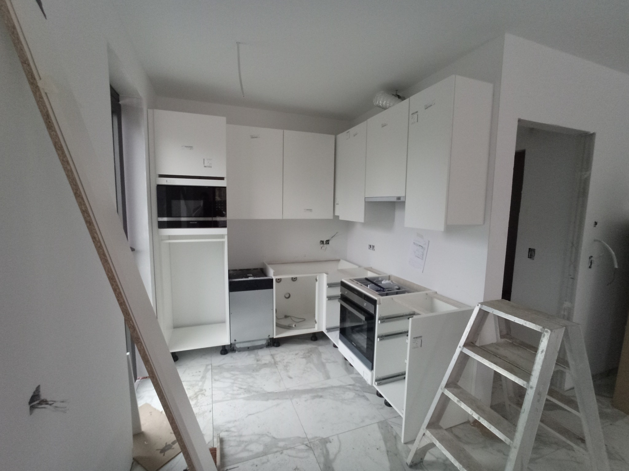 Montaż białej kuchni: widok na niedokończoną zabudowę z metalową drabinką, płytą grzewczą i piekarnikiem w tle, na marmurowej podłodze.