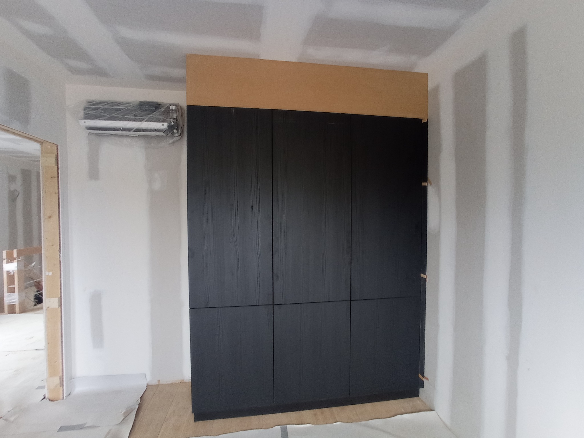 Ciemna, sześciodrzwiowa szafa wnękowa z drewnianymi drzwiami i jasnym panelem górnym, zamontowana w pomieszczeniu w trakcie remontu, z klimatyzatorem zabezpieczonym folią.