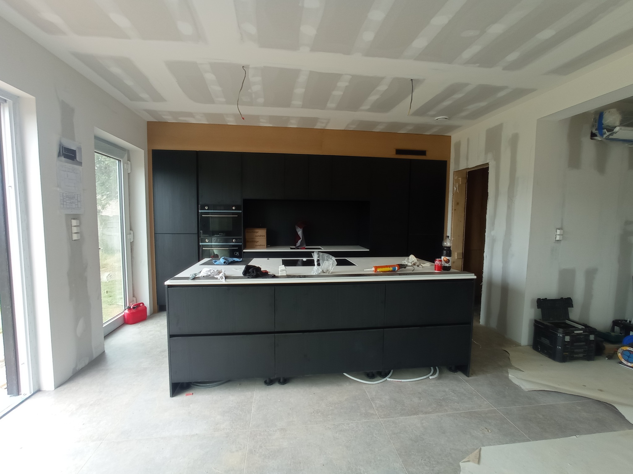 Ciemna zabudowa kuchenna z wyspą i białym blatem w surowym wnętrzu, widoczne narzędzia i materiały budowlane, sufit w trakcie wykończenia płytami gipsowymi.