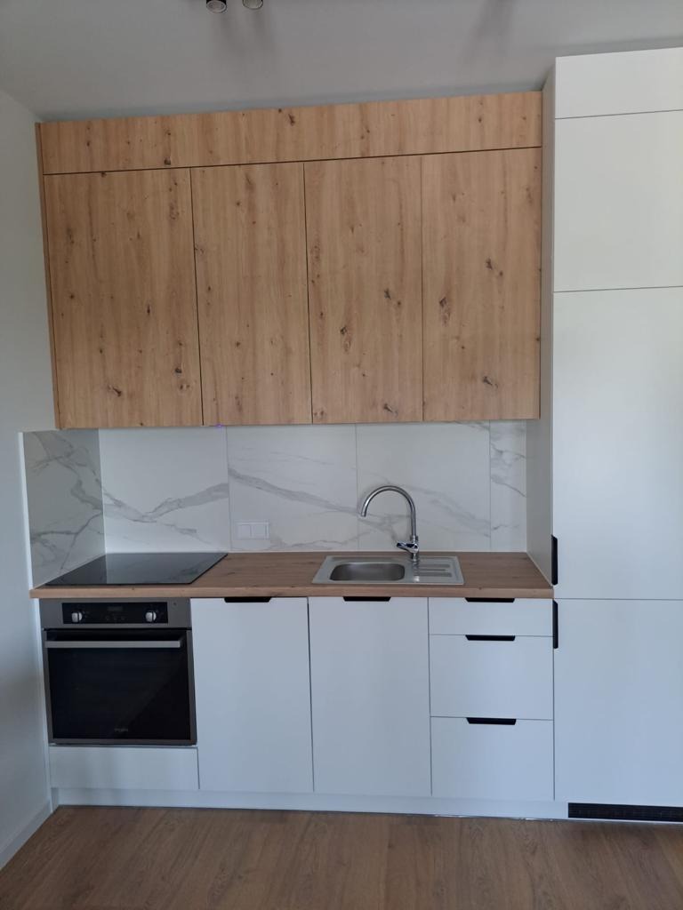 Nowoczesna kuchnia z białymi frontami szafek dolnych i drewnopodobnymi górnymi, marmurowe płytki nad blatem, płyta indukcyjna i zlew ze stali nierdzewnej.