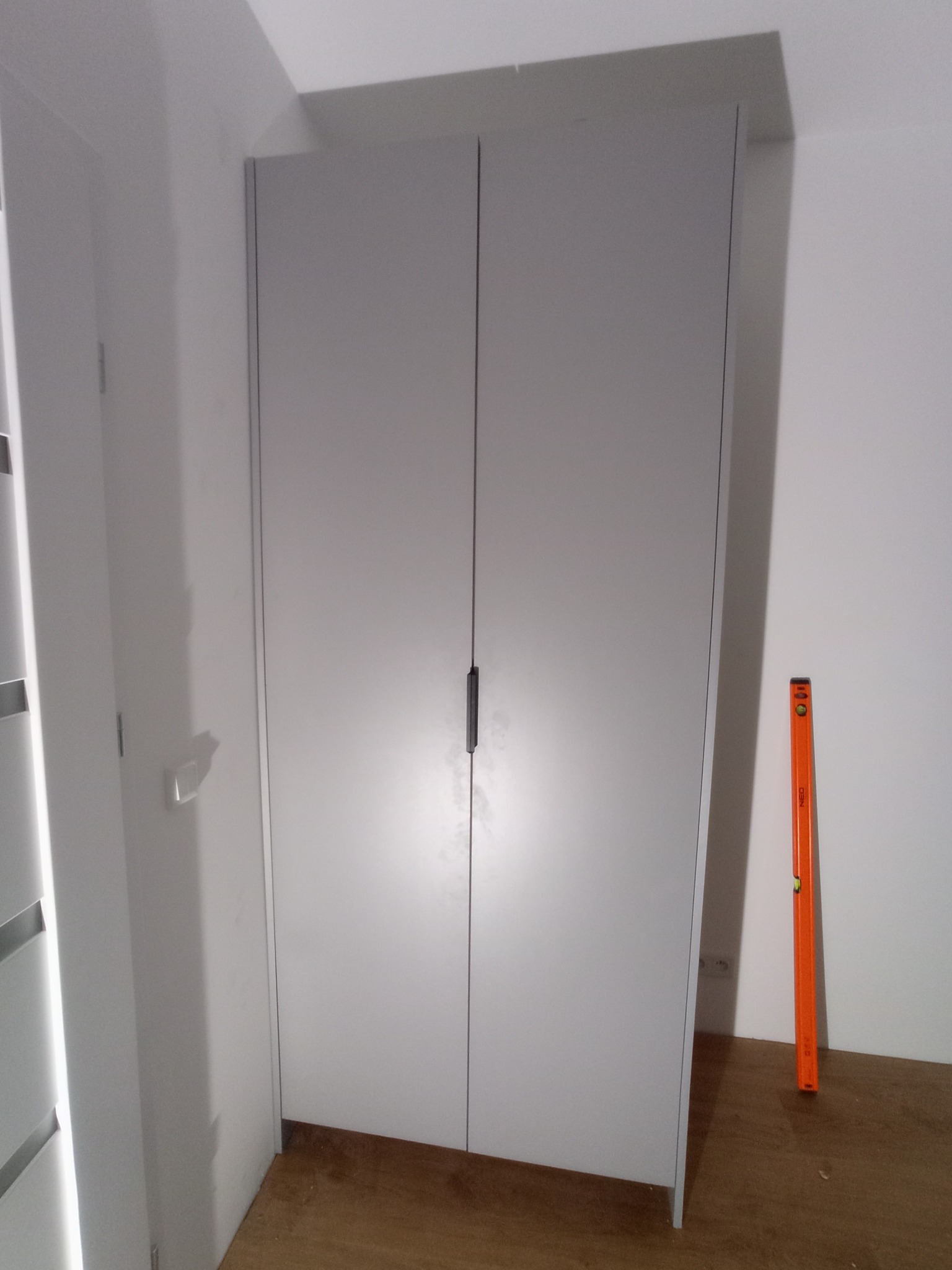 Szara szafa dwudrzwiowa z długim, czarnym uchwytem, stojąca w rogu pokoju na drewnianej podłodze, z pomarańczową poziomicą opartą o bok szafy.