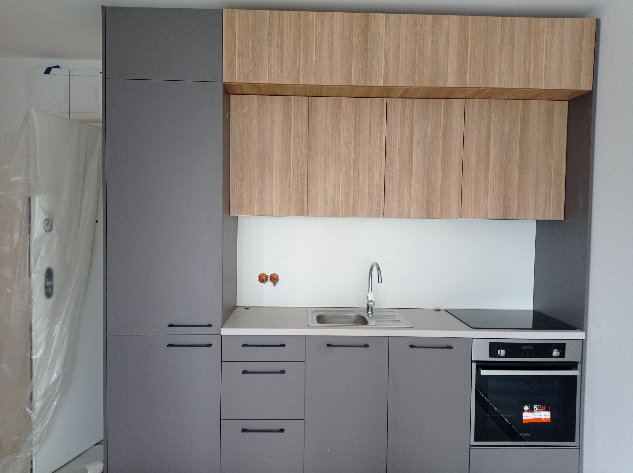 Nowoczesna zabudowa kuchenna w odcieniach szarości i drewna, z widocznym zlewem, płytą indukcyjną i piekarnikiem, częściowo zasłonięta folią ochronną podczas montażu.