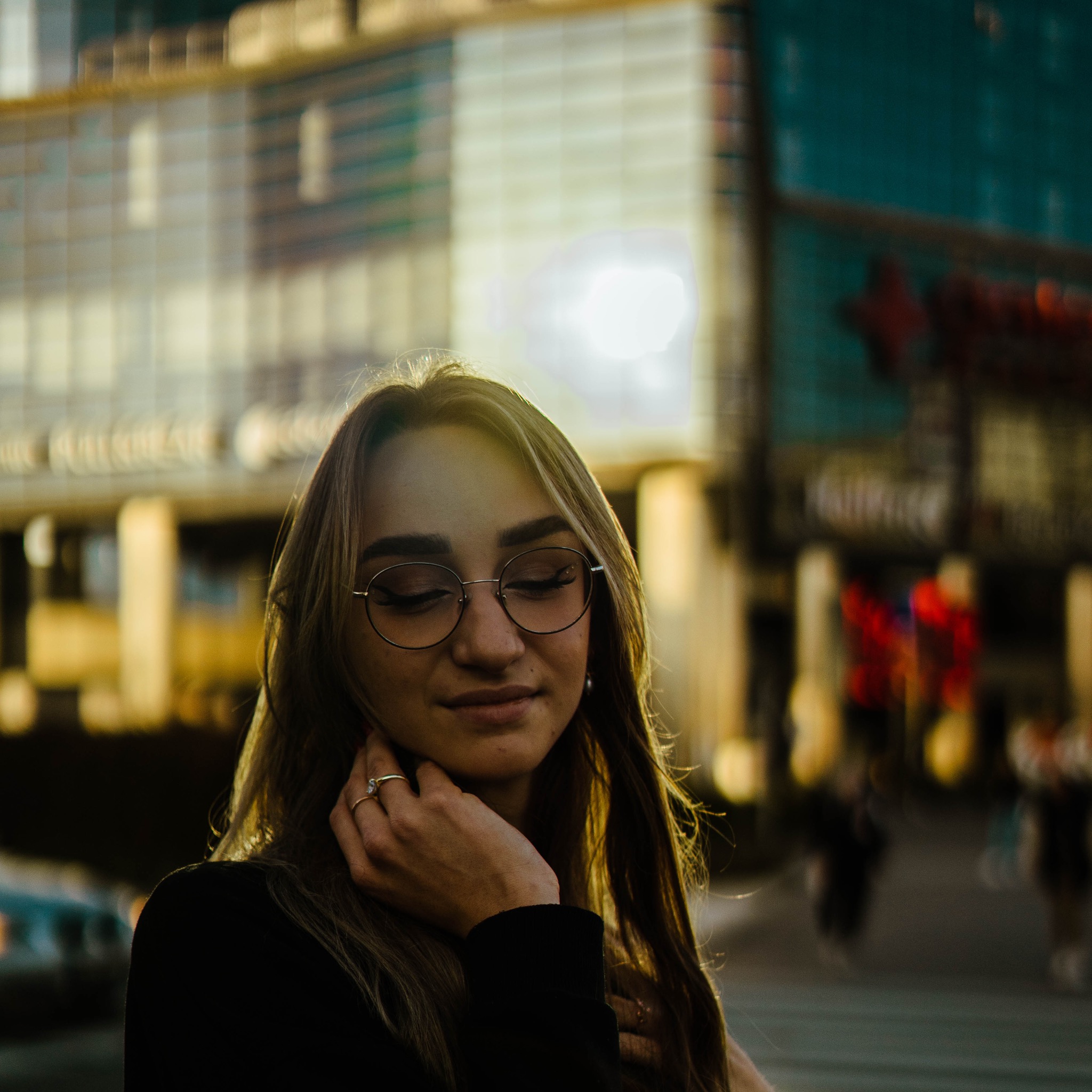 Portret kobiety w okularach, z ręką przy szyi, na tle miejskiej zabudowy z refleksami zachodzącego słońca.