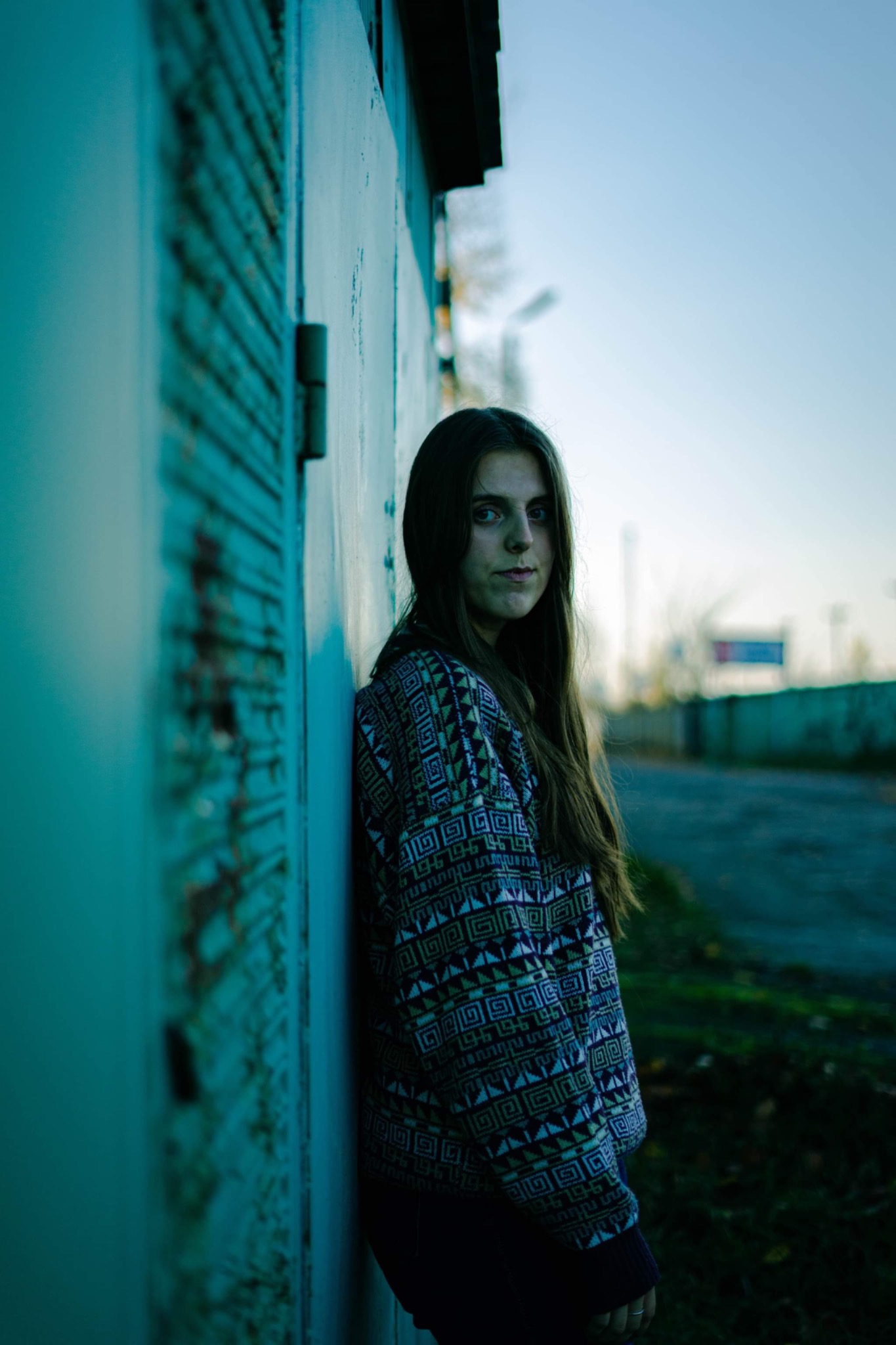 Portret młodej kobiety opierającej się o turkusową, zardzewiałą ścianę w swetrze z geometrycznym wzorem, w tle widoczne drzewa i niebieskie niebo.