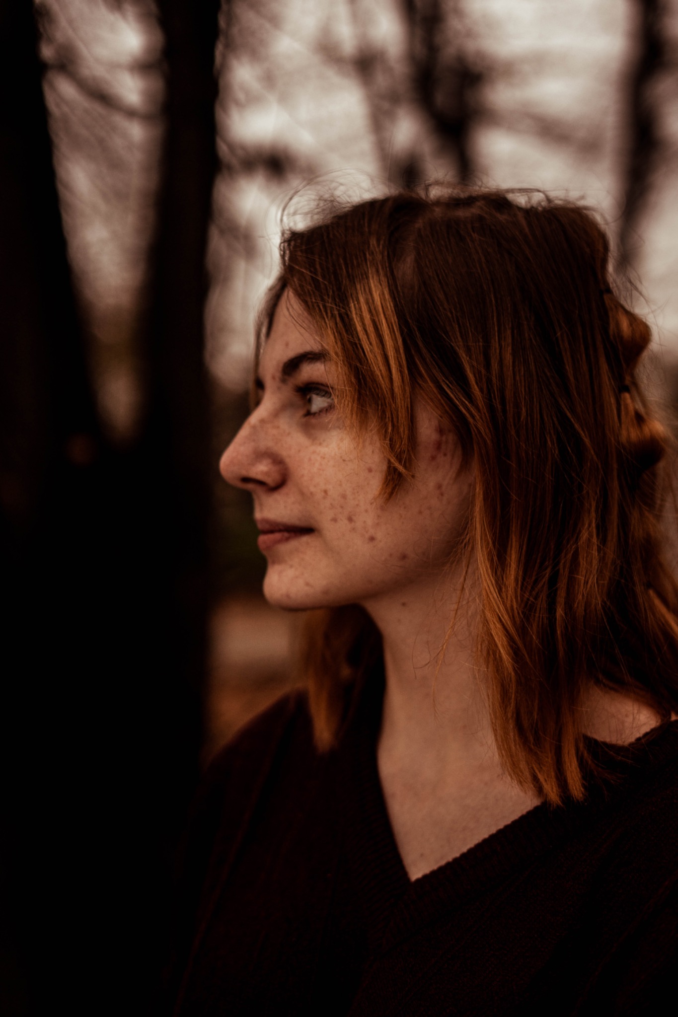 Portret z profilu młodej kobiety z rudymi włosami i piegami, zamyślony wyraz twarzy, ciemne tło z zamazanymi drzewami.