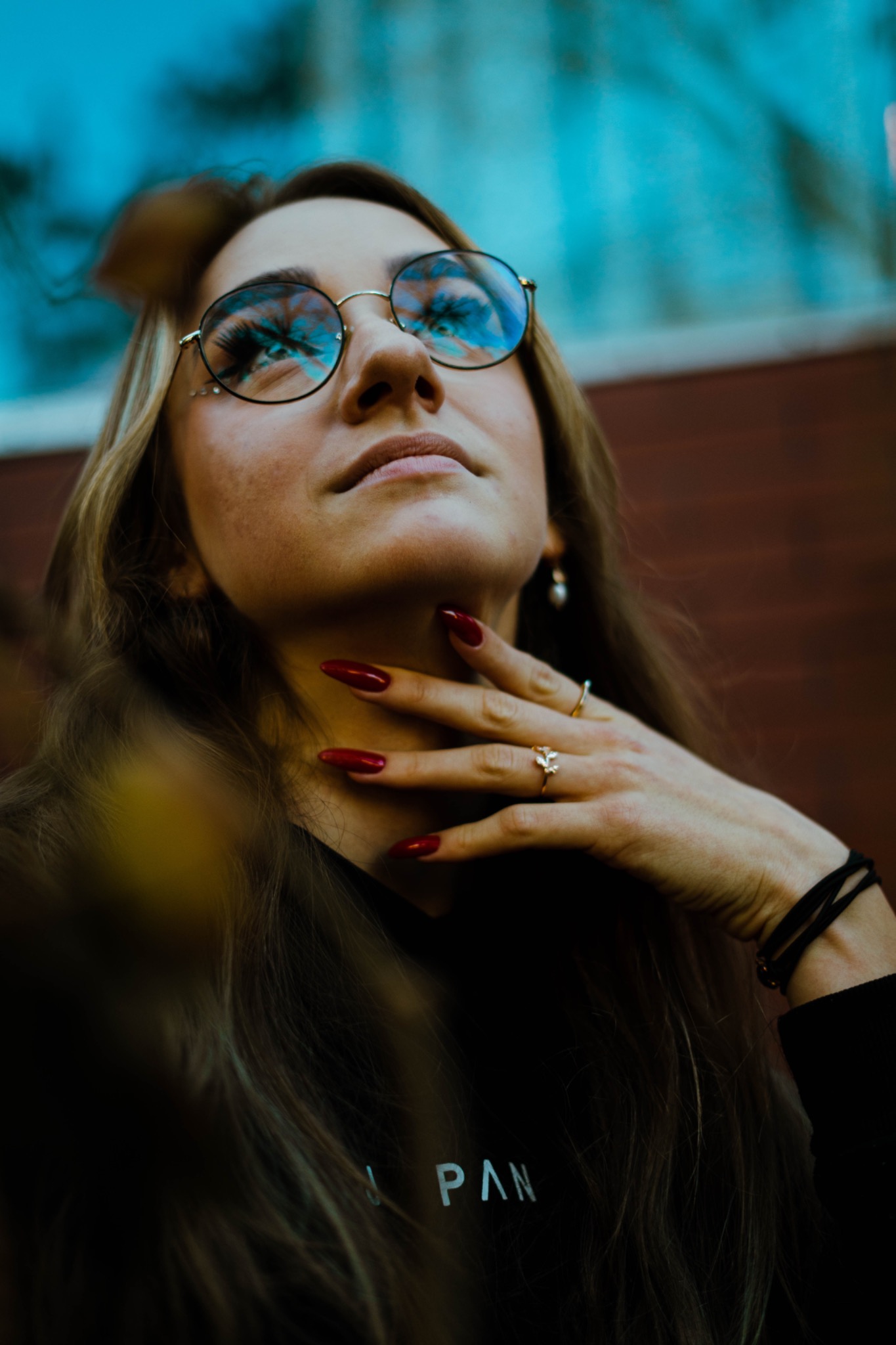 Portret kobiety z uniesioną głową, noszącej okrągłe okulary z odbijającym się niebem, czerwonymi paznokciami i delikatną biżuterią.