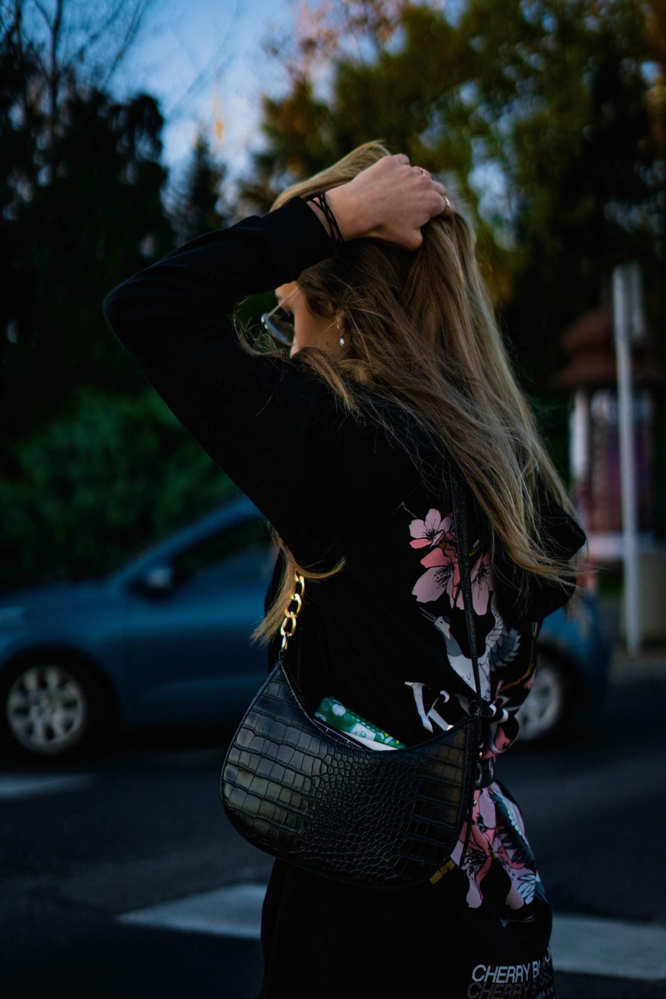 Kobieta odwrócona tyłem, poprawiająca włosy, ubrana w czarną bluzę z motywem kwiatowym i dźwigająca czarną torebkę na złotym łańcuszku na tle ulicy i samochodu.