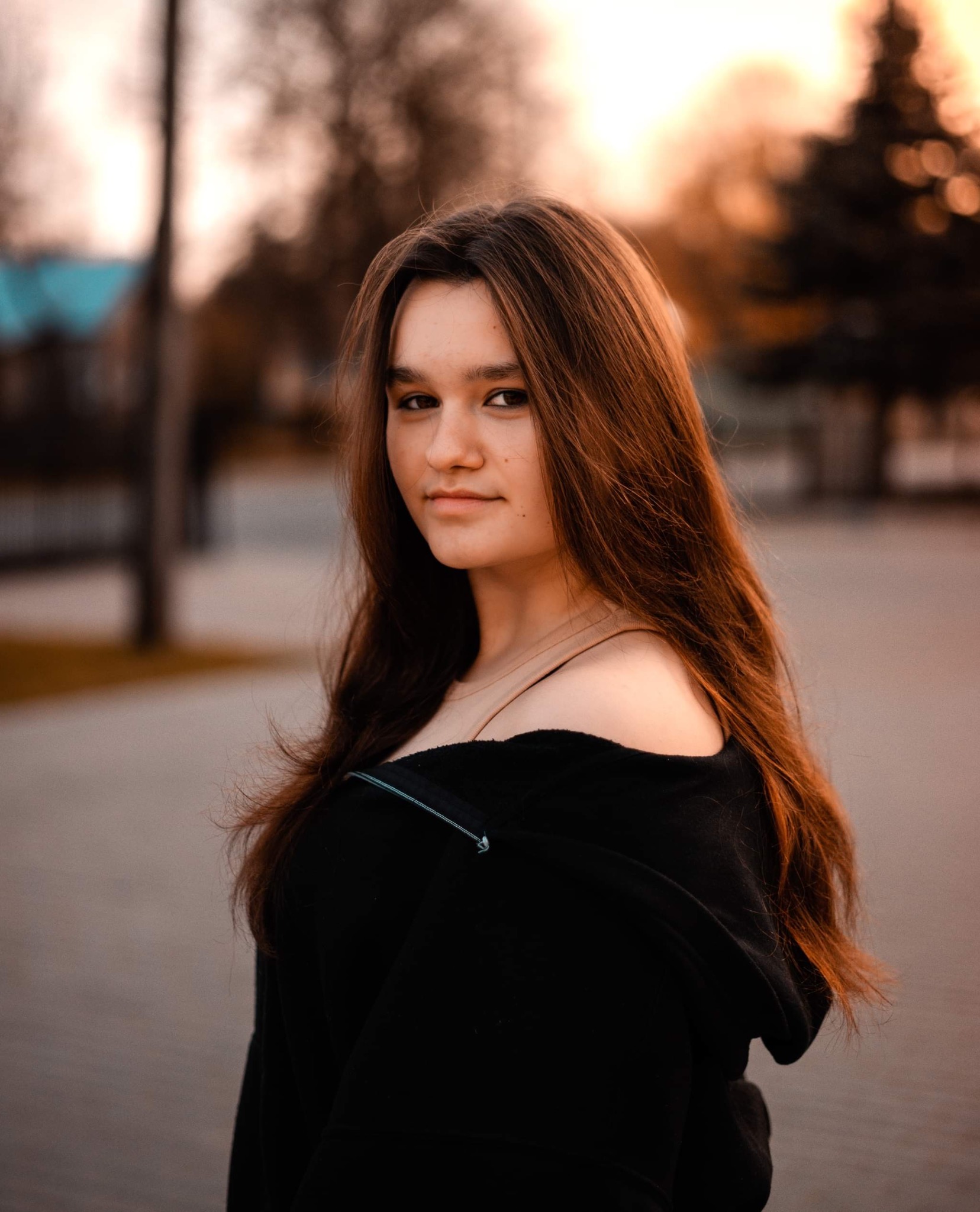 Portret młodej kobiety z długimi, brązowymi włosami, ubrana w czarną bluzę z odkrytymi ramionami, na tle rozmytego parku o zachodzie słońca.