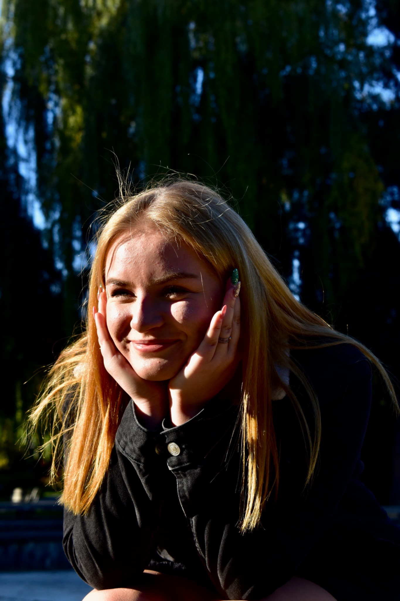 Portret uśmiechniętej blondynki w czarnej kurtce, z rękami podpartymi pod brodą, na tle rozmytej zieleni drzew