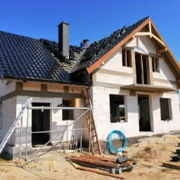 Montaż czarnej dachówki na nowym domu w trakcie budowy; widoczny dekarz pracujący na dachu, komin, surowa konstrukcja budynku z pustaków oraz drabiny. Częściowo pokryty dach, błękitne niebo w tle.