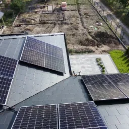 Instalacja paneli słonecznych na dachu z szarej dachówki ceramicznej, widok z góry na posesję z ogrodem w trakcie aranżacji.