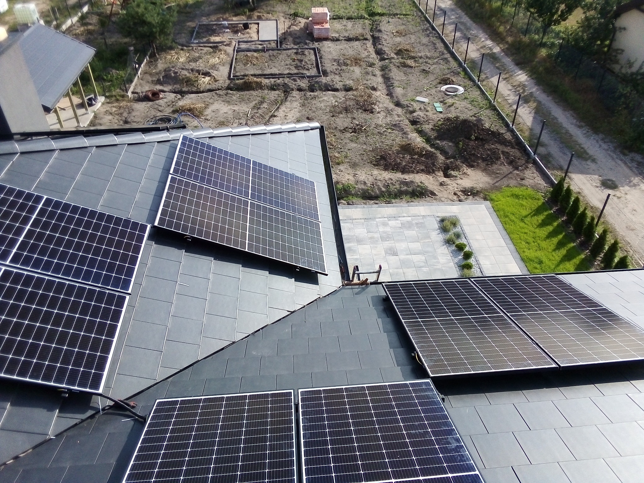 Instalacja paneli słonecznych na dachu z szarej dachówki ceramicznej, widok z góry na posesję z ogrodem w trakcie aranżacji.