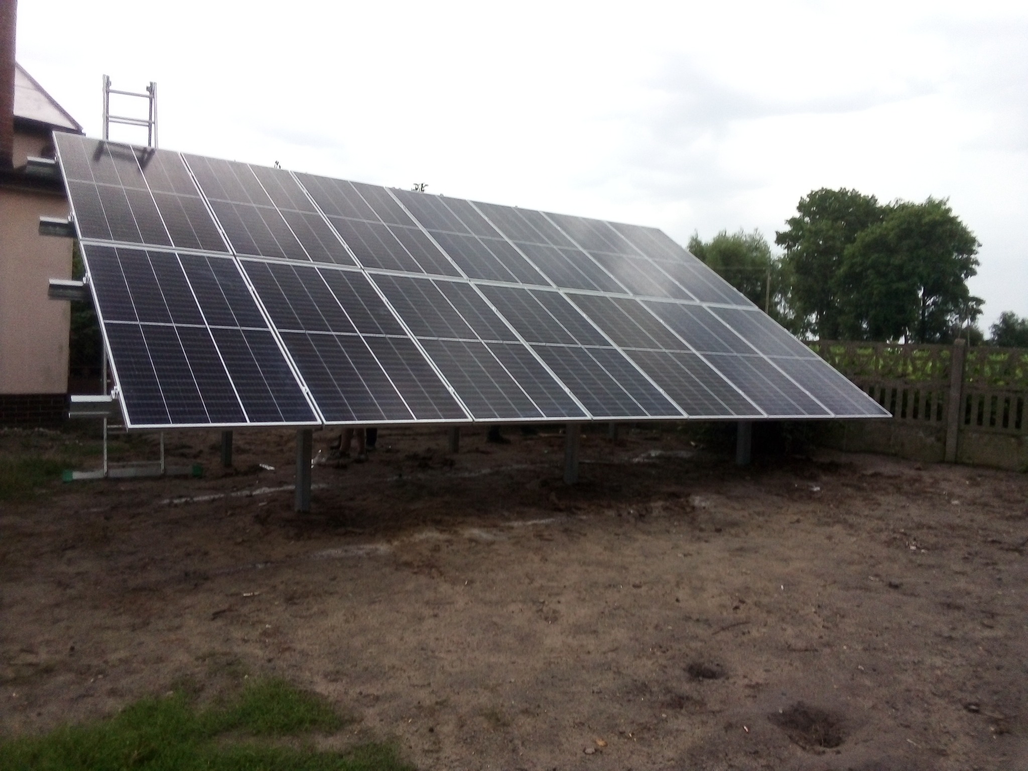 Instalacja paneli słonecznych na konstrukcji wsporczej, widok z poziomu gruntu na tle pochmurnego nieba i ogrodzenia.