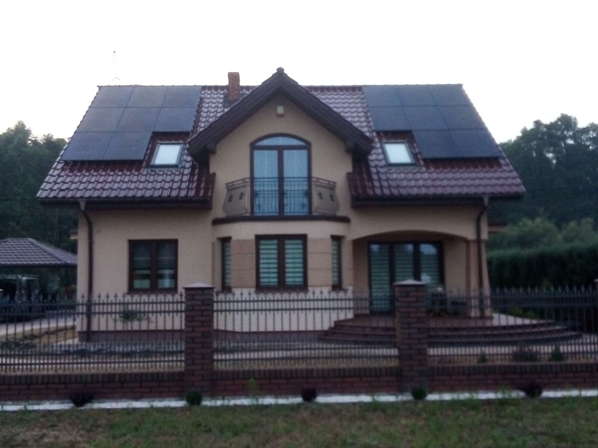 Dwukondygnacyjny dom jednorodzinny z panelami słonecznymi na dachu z czerwonej dachówki, widok z zewnątrz na ogrodzenie z ceglanymi słupkami.