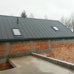 Nowy, grafitowy dach z blachy trapezowej z dwoma oknami dachowymi i kominem wentylacyjnym na budynku w trakcie budowy, z widoczną surową ścianą z cegły.