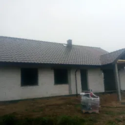 Dom w stanie surowym zamkniętym w Mąkoszycach, z szarą dach&oacute;wką ceramiczną i widocznym kominem, sfotografowany w mglisty dzień.