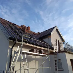 Pracownik w trakcie układania dachówki na skośnym dachu domu jednorodzinnego, widoczne rusztowanie i zapas dachówek na podeście.