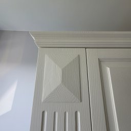 Woodii Damian Mrzygłód - Fragment białej szafy z frezowanymi drzwiami i ozdobnym zwieńczeniem, widoczna struktura drewna.