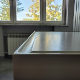 Woodii Damian Mrzygłód - Fragment blatu w jasnym odcieniu z widoczną strukturą drewna, w tle okno z widokiem na zieleń i grzejnik.