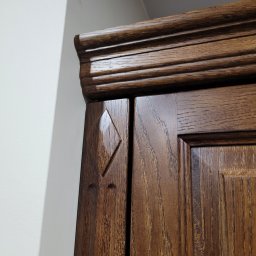 Woodii Damian Mrzygłód - Fragment górnej części ciemnobrązowej drewnianej szafy z dekoracyjnym frezem i romboidalnym elementem na drzwiach, przylegającej do białej ściany.