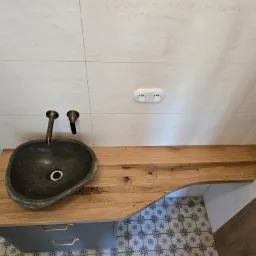 Łazienka z blatem drewnianym i kamienną umywalką. Widoczne białe płytki na ścianie z notatkami i gniazdko elektryczne. Ujęcie z góry, prezentujące unikalny design.