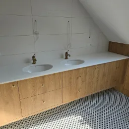 Łazienka na poddaszu z drewnianą szafką pod umywalki i białym blatem, złote krany, wzorzysta podłoga. Widoczne przygotowania do montażu oświetlenia.