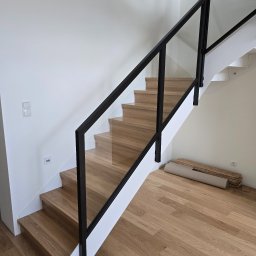 Woodii Damian Mrzygłód - Drewniane schody z czarną balustradą i szklanym wypełnieniem. Na podłodze zwój i deski podłogowe. Widok z dołu na nowoczesne schody w jasnym wnętrzu.